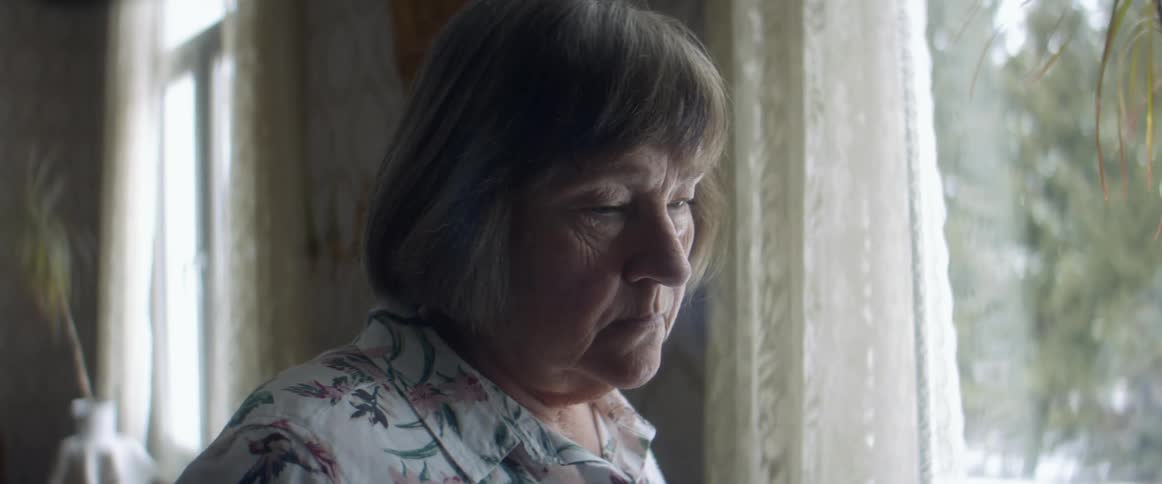 An old woman standing by a window in a bedroom looking down sadly