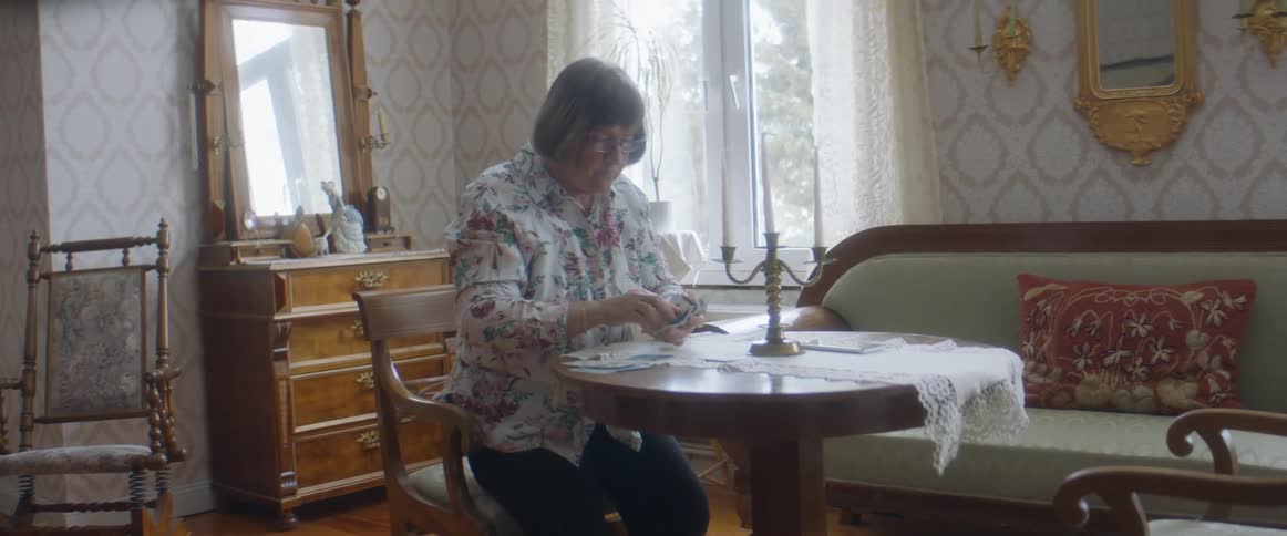 A woman in a room sitting on a chair at a table with a wall and window in the background