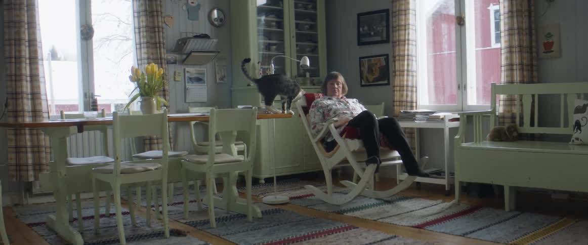 A woman in a room sitting on a rocking chair with a cat on a table next to her
