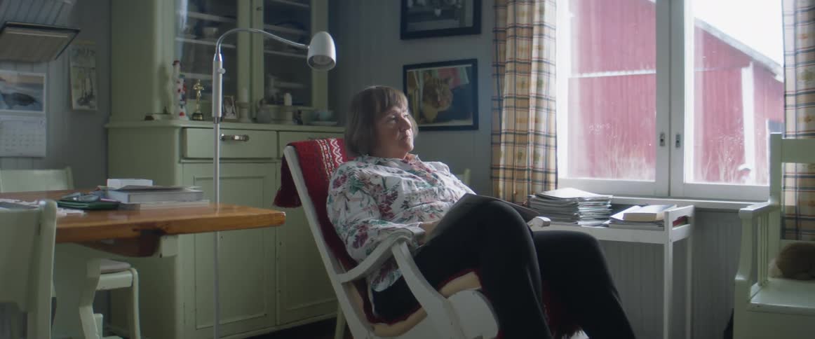 A woman sitting in a rocking chair and taking off her glasses with a wall and window in the background