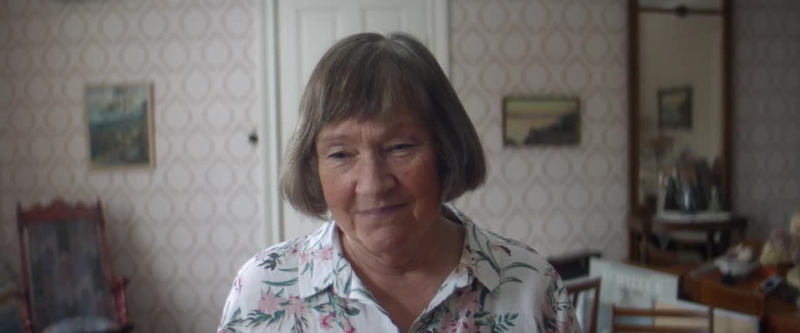An elderly woman walks towards the camera in a house before stopping and smiling