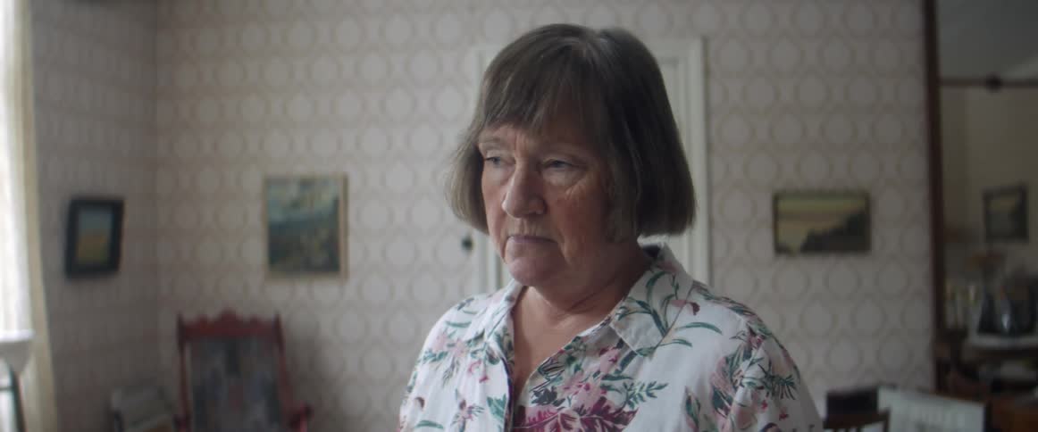 An elderly woman stands indoors and looks towards the window with a look of boredom on her face