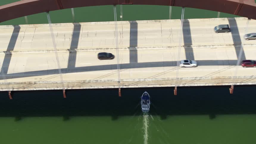 An aerial view of a boat driving under a bridge with cars passing overhead