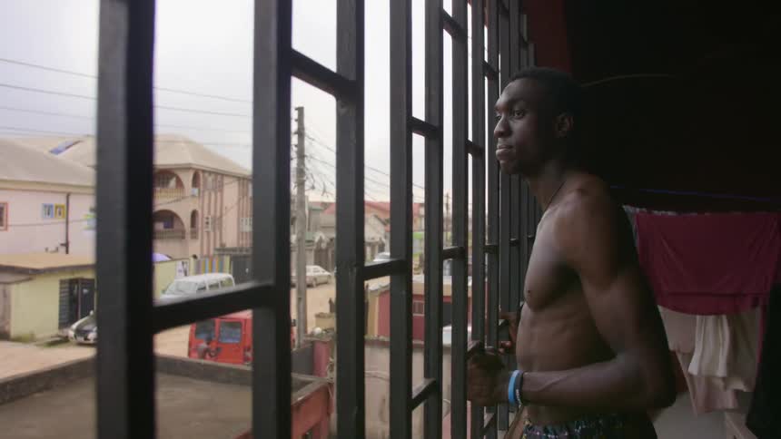 A man looks out a window to the quiet street with parked cars