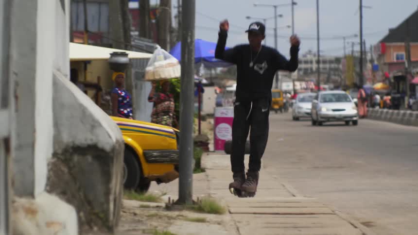A teenager rollerskates down a street with cars driving on and then jumps on to a wall and back on to the street