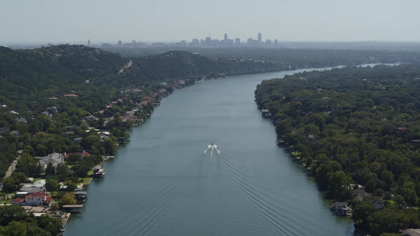 Boat moves along river between forest shores pasts houses with foggy city on horizon
