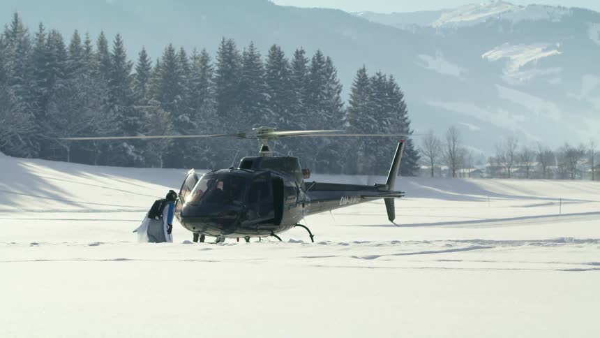 A person in a wing suit walks towards a helicopter waiting in a snowy field during the day