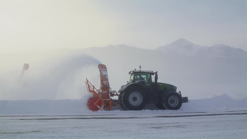 A tractor with a snow remover with a mountain range the sky and clouds in the background