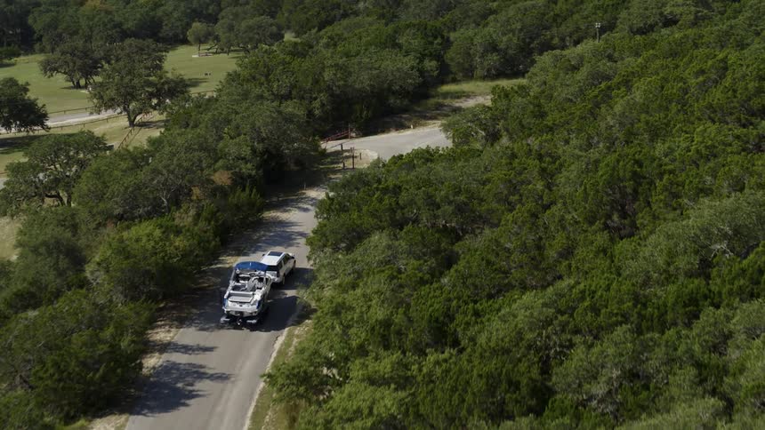 A vehicle pulls a boat on a trailer along behind it as it travels down a road through a woodland during the day