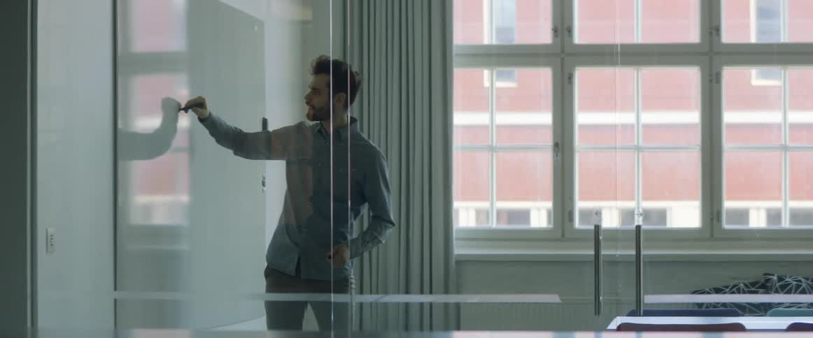 A man writes on a white board in an office as he talks