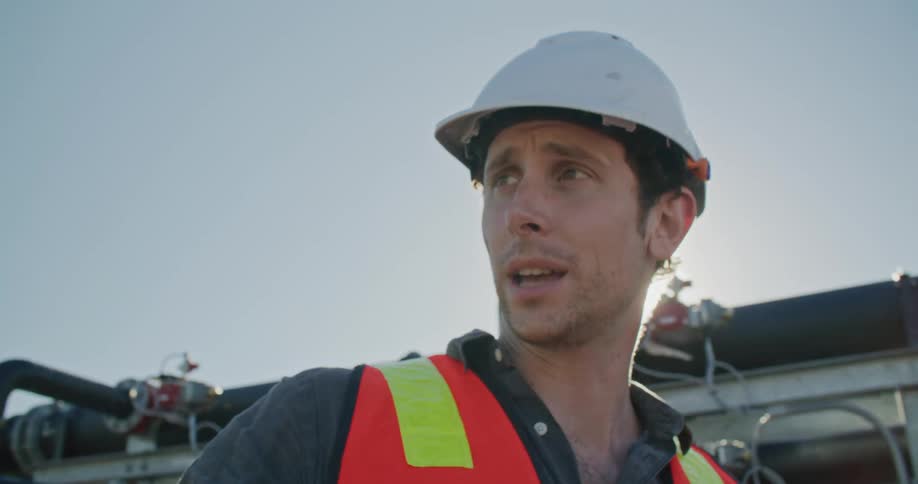 Two men with helmets stand and talk next to large machinery