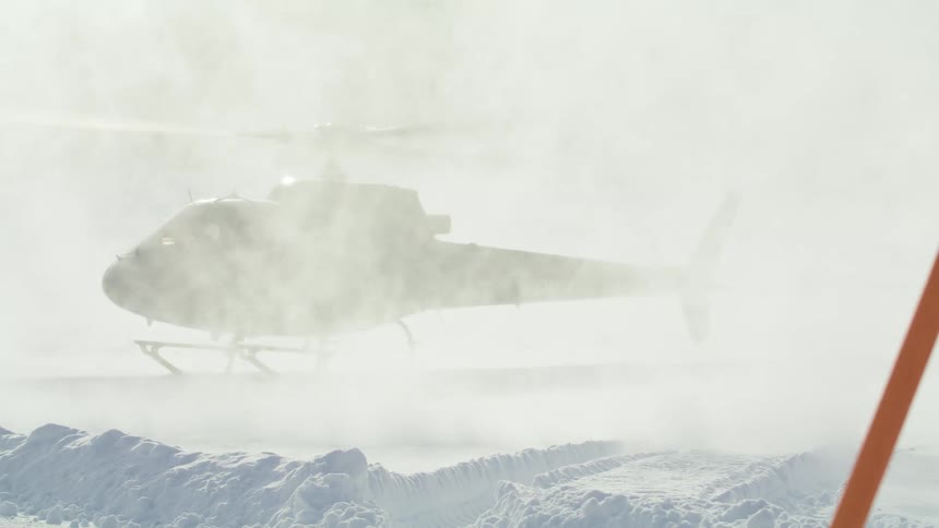 Snow flies up as a helicopter lands at the base of a mountain