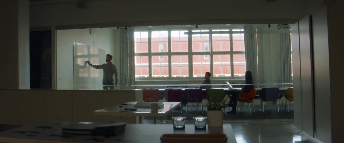 A man standing in a room writing on a whiteboard with another man and woman sitting on chairs by a conference table
