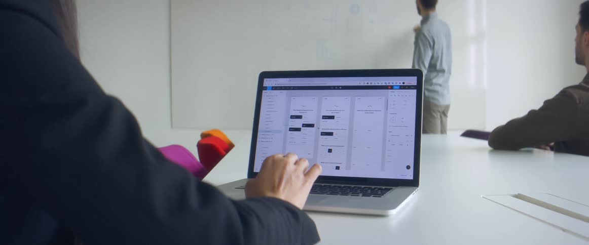 Office workers sit at table with laptop while another gives a presentation on a whiteboard
