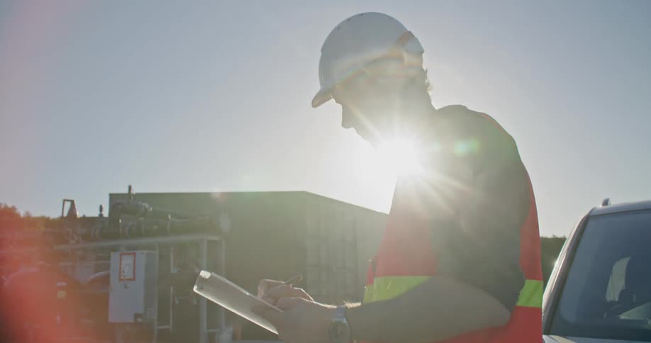 A flare of sunlight hitting a man with a clipboard leaned against a car outside at a worksite taking notes