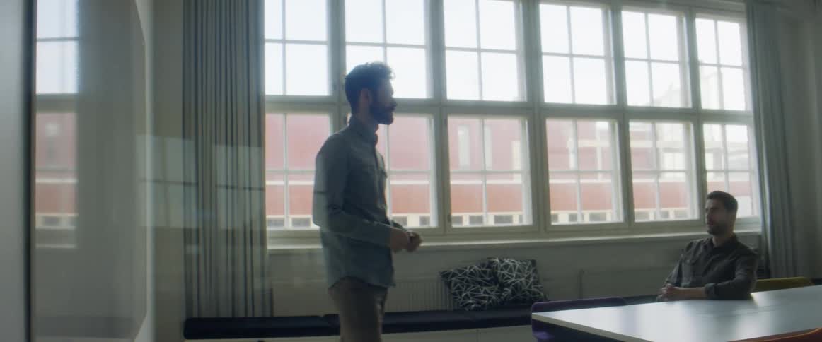 A man stands in front of a large table and a man and woman in an office