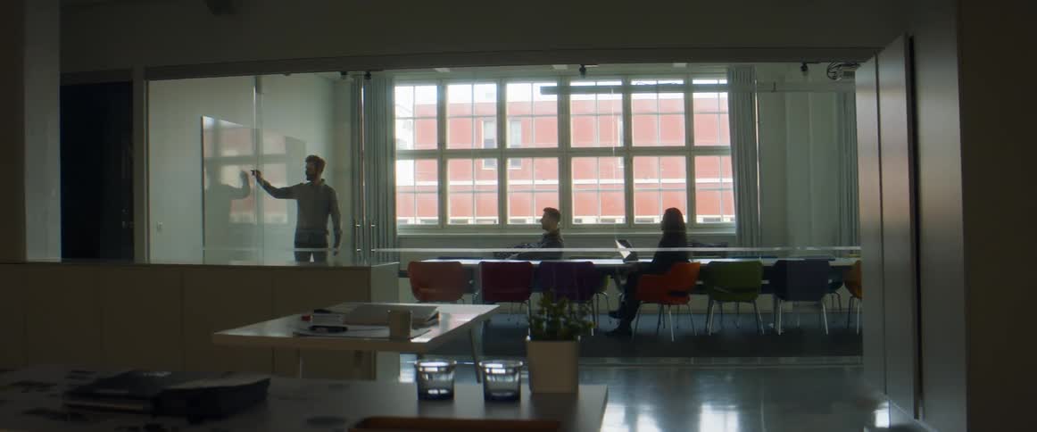 A man stands in a large office room talking to two other people and writing on a whiteboard