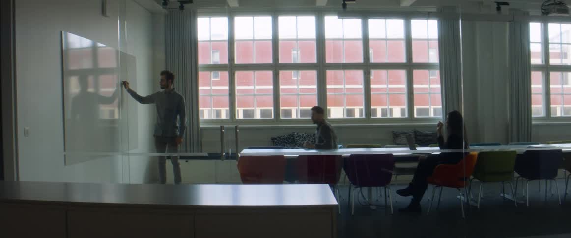 A man stands in front of two people in a large office room and writes on a whiteboard