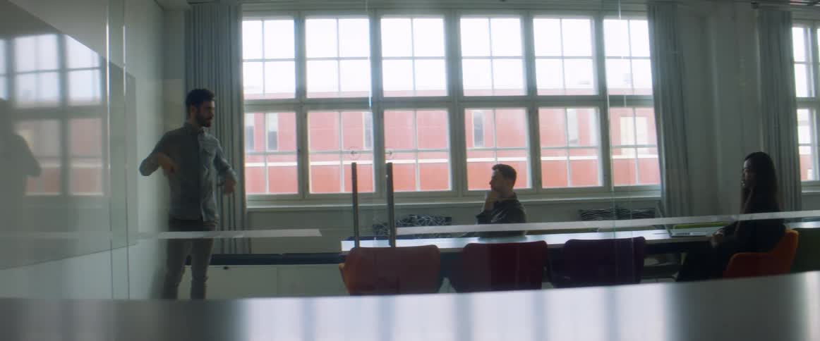 A man writes on a whiteboard in front of two people in a large office room