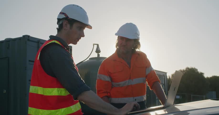 Two construction workers view a laptop thats placed on the hood of a vehicle at a construction site