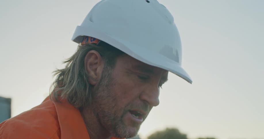 A man with a hardhat talking and pointing at a laptop with trees the sky and clouds in the background