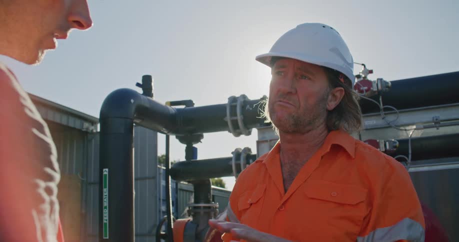 Two men with hardhats talking with pipes the sky and clouds in the background
