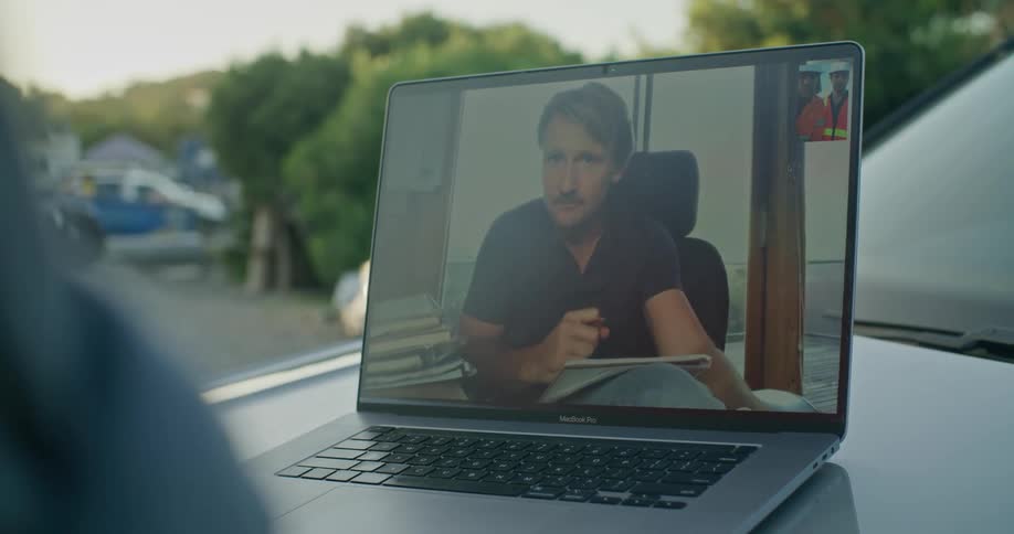 An engineer discusses blueprints with his colleagues during a video call on a laptop placed on the hood of his car at a site