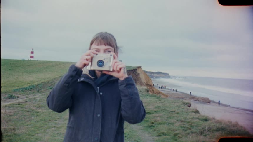 A woman with a coat and a camera taking a picture with the ocean sky and clouds in the background on analog Film