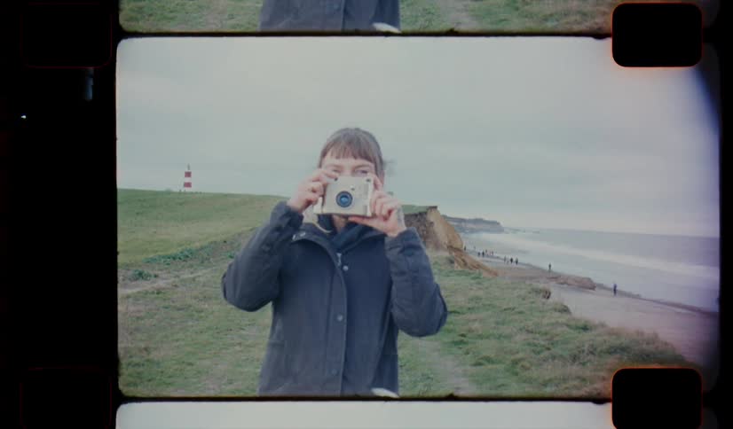 A woman wearing a coat stands near the coast while holding up a camera on analog Film