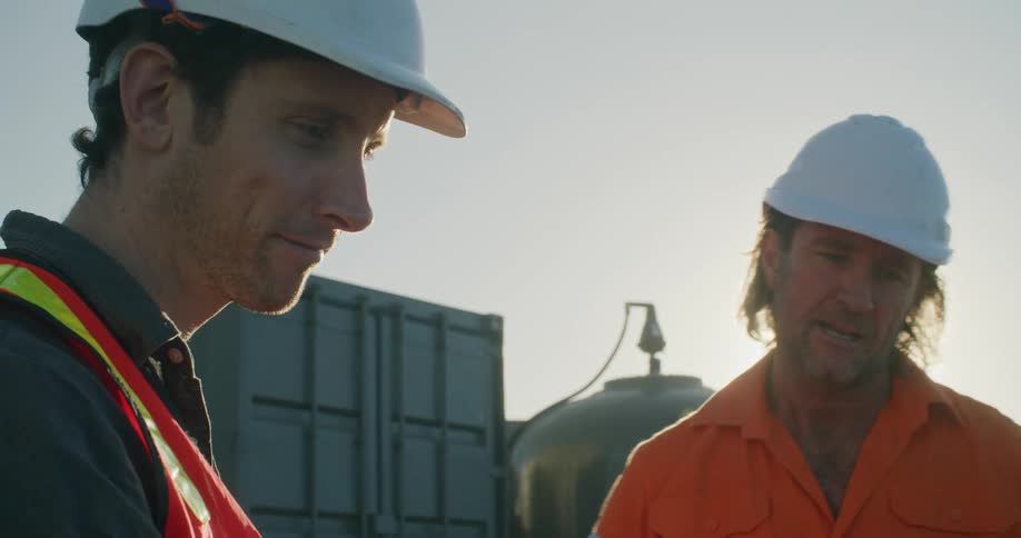 A pair of workers in personal protective equipment speak with one another while consulting a laptop resting on the hold of a car outdoors during the day