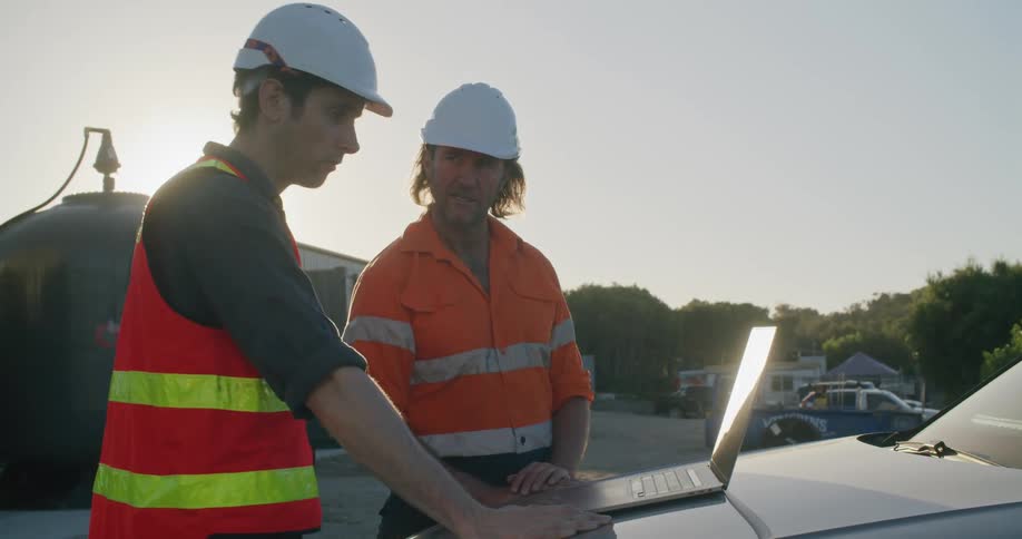 Two construction workers stand beside each other other side talking by a laptop