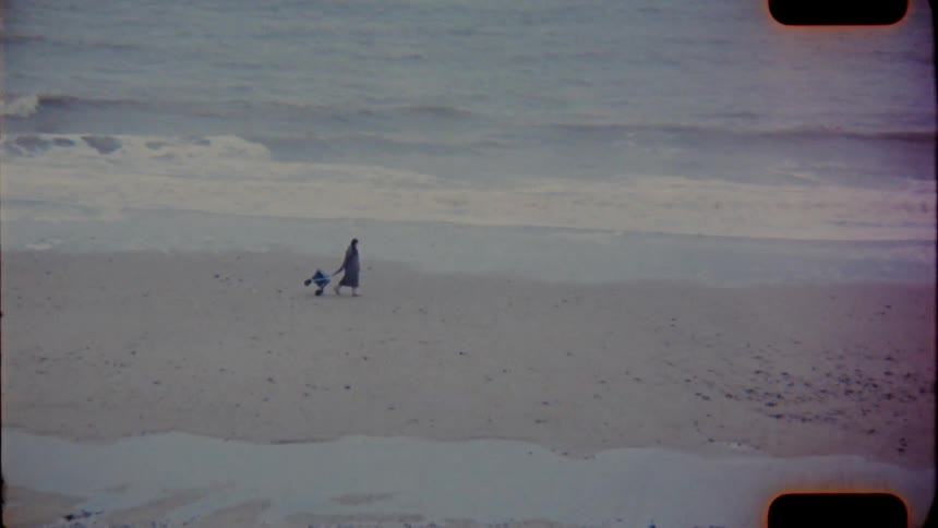 A woman pulling a wheeled cart behind her walks along a sandy beach as waves wash ashore on analog Film