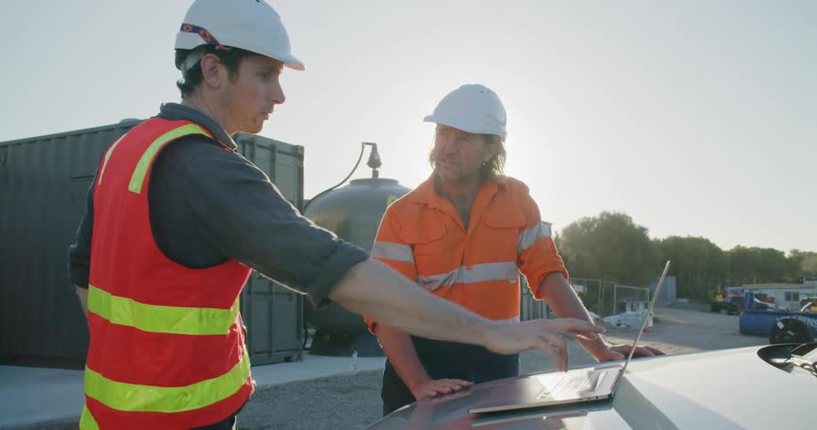 Two construction workers stand outside beside a laptop talking together