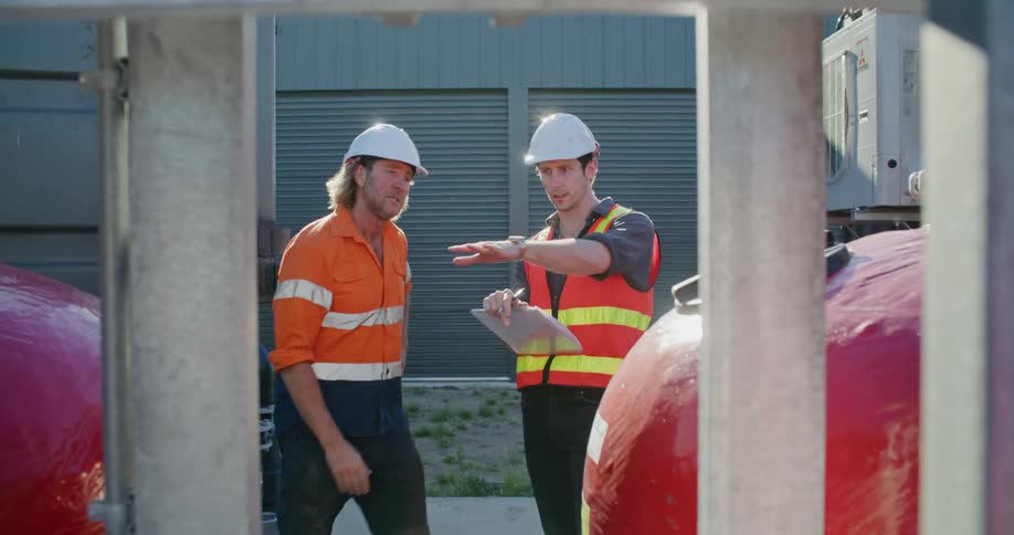Construction supervisors wearing white hard hats talking in front of red container tanks