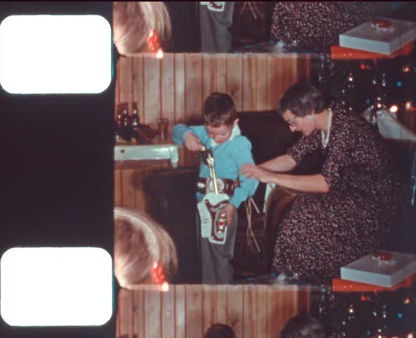 A woman helps a boy tie a toy gun holster around his waist in front of a christmas tree