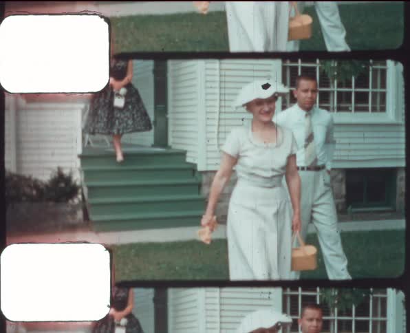 A woman with a hat and a bag walking with a house in the background