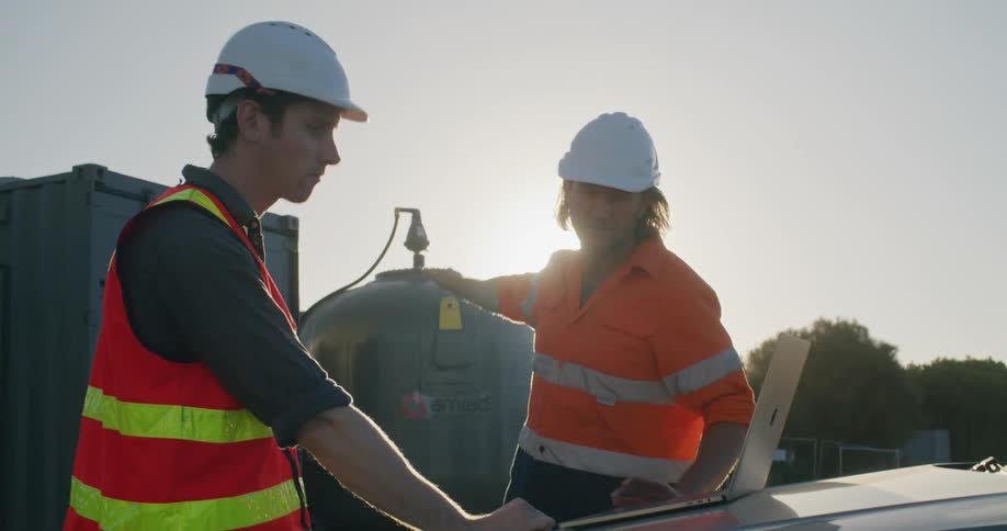 Two men with hardhats looking at a laptop with trees the sky and clouds in the background