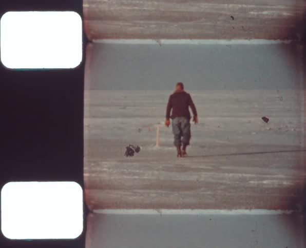 Man walks across snowy plain toward short staff stuck upright in ground