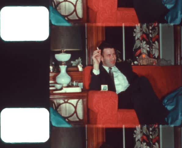 A white male sitting on a couch smoking a cigarette being filmed on vintage film