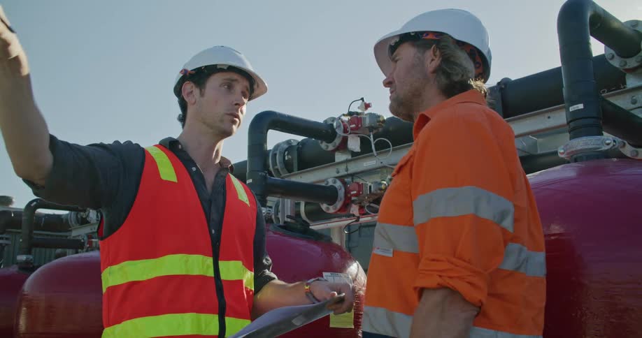 Two men with hard hats on stand outside and talk as one points off in to the distance