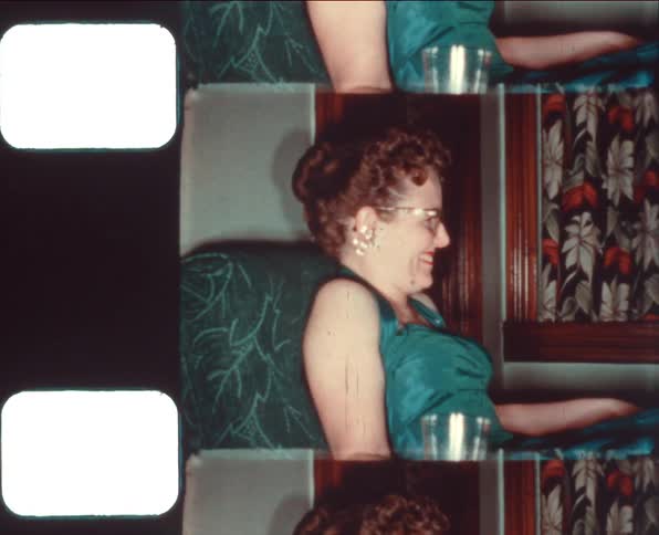 A smiling woman sticks her tongue out at the camera as she drinks and speaks while sitting on a chair in an evening dress