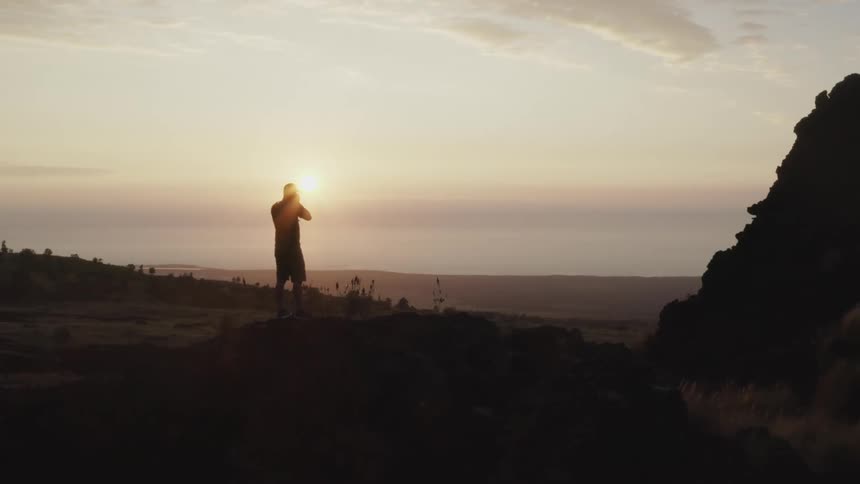 A photographer climbs atop a rock and uses a camera to take a photograph of the countryside in the evening
