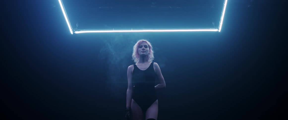 A woman with short blonde hair walks seductively forward while illuminated by overhead neon lights