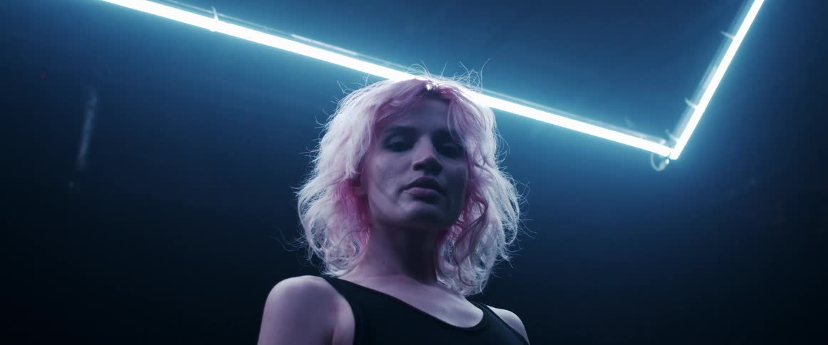 A woman with short blond hair stands under a neon light while staring directly into the camera