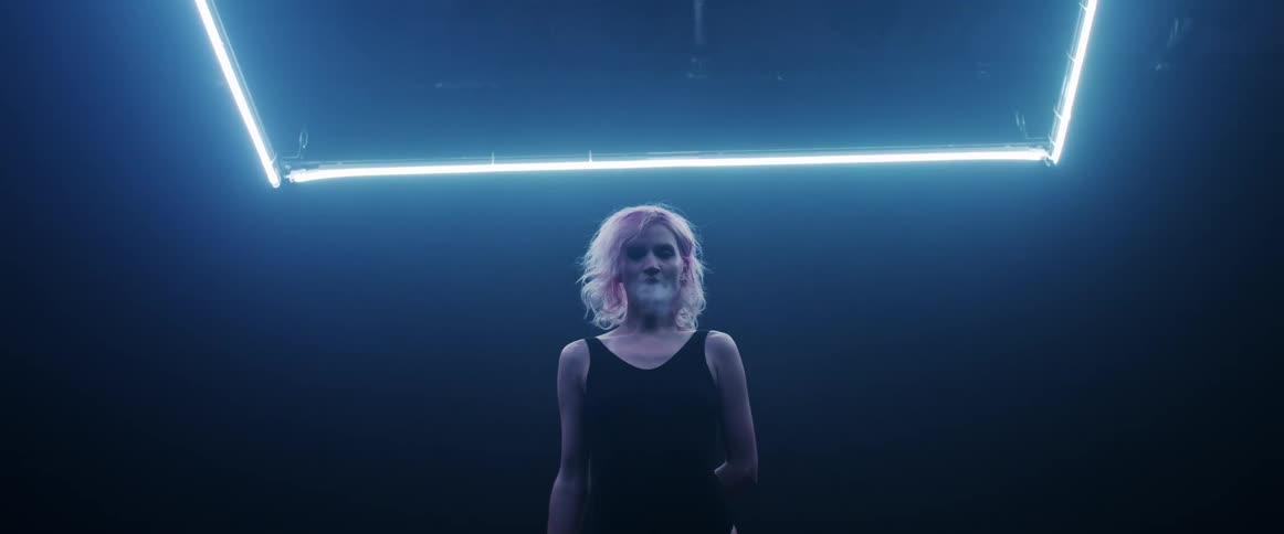 A woman stands in a dark room under a neon light as she exhales smoke