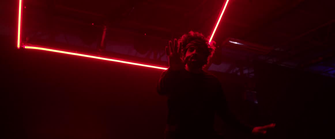 A man with puffy hair and a beard dances indoors beneath red neon lights