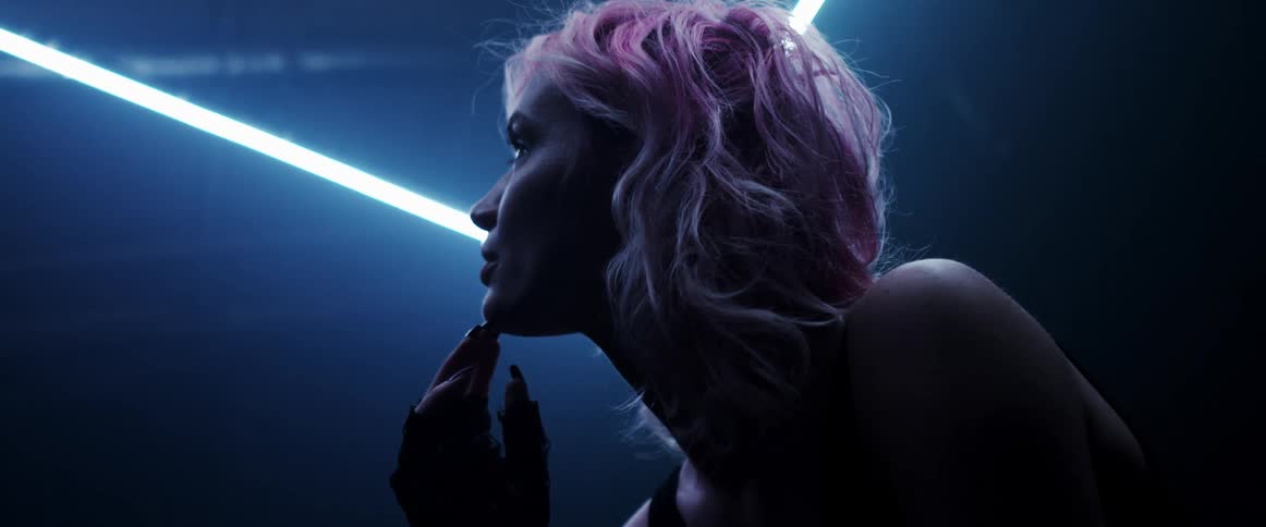 Blonde woman with pink highlights looks up at bright neon lights in dark room
