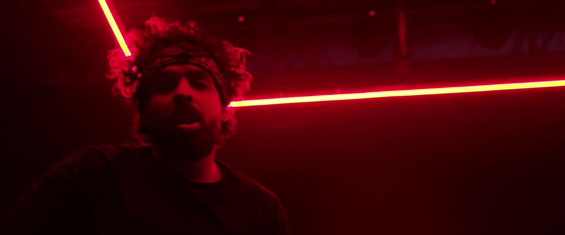 A man standing in a room with red lighting performs a dance move with his hands while mouthing words