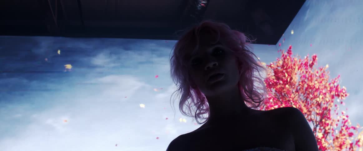 A woman in sparkling dress looks into the camera while standing in front of a video wall in a dark room
