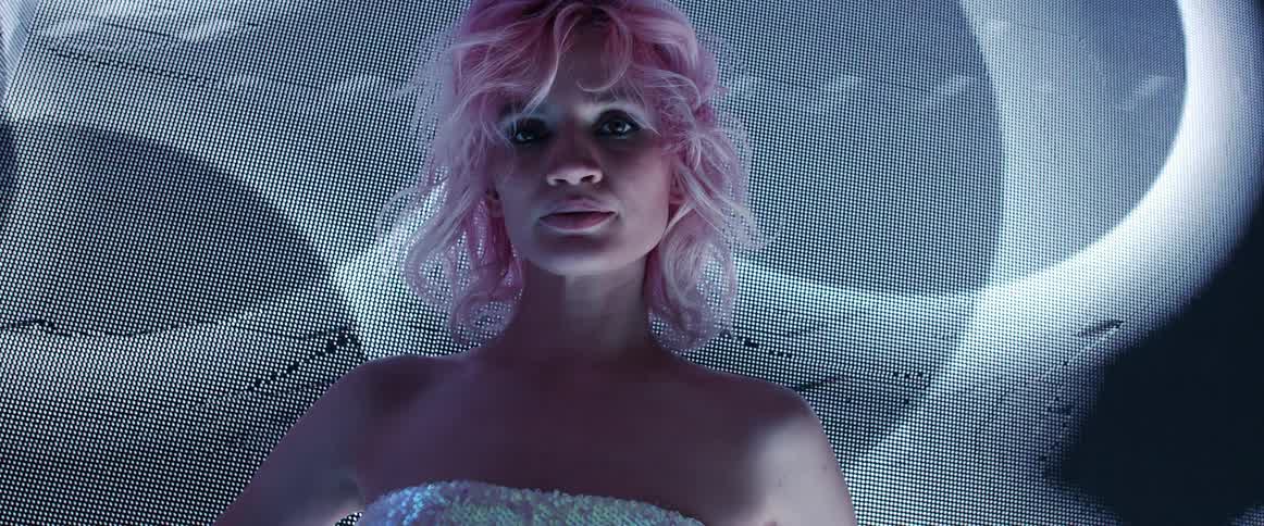 A woman with short blond hair stands in front of a huge screen displaying abstract imagery